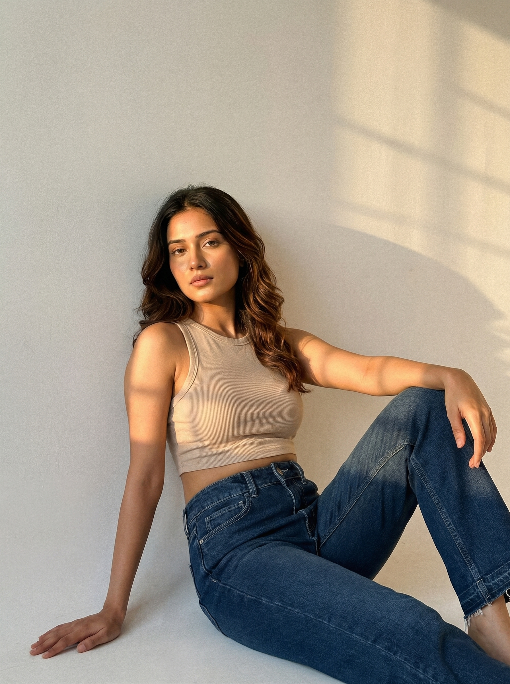 Woman sitting against a white wall wearing a beige tank top and NOIR Core indigo jeans.