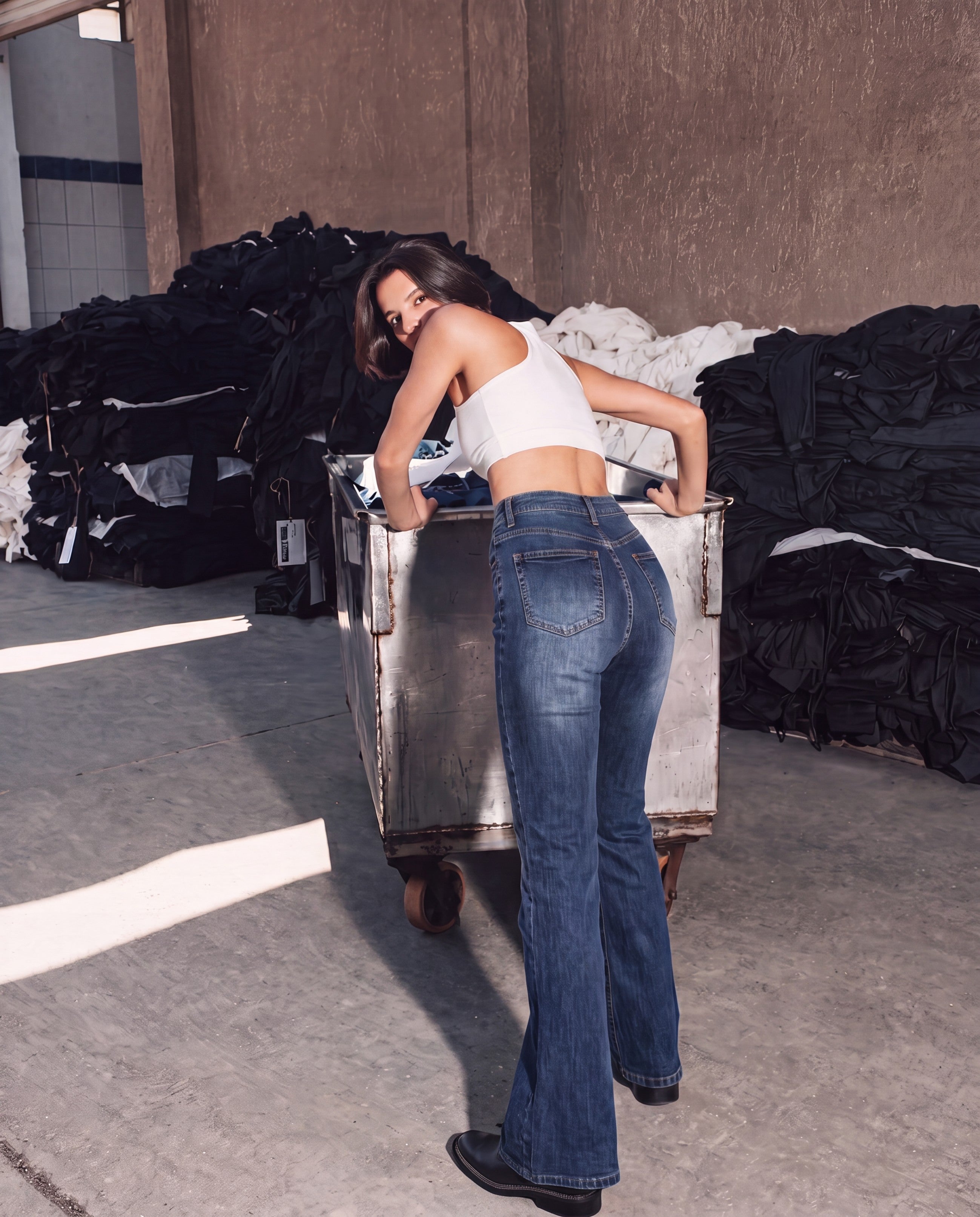 A woman wearing blue flared jeans and a white crop top leans over a metal cart in an industrial warehouse setting with piles of folded clothing in the background.