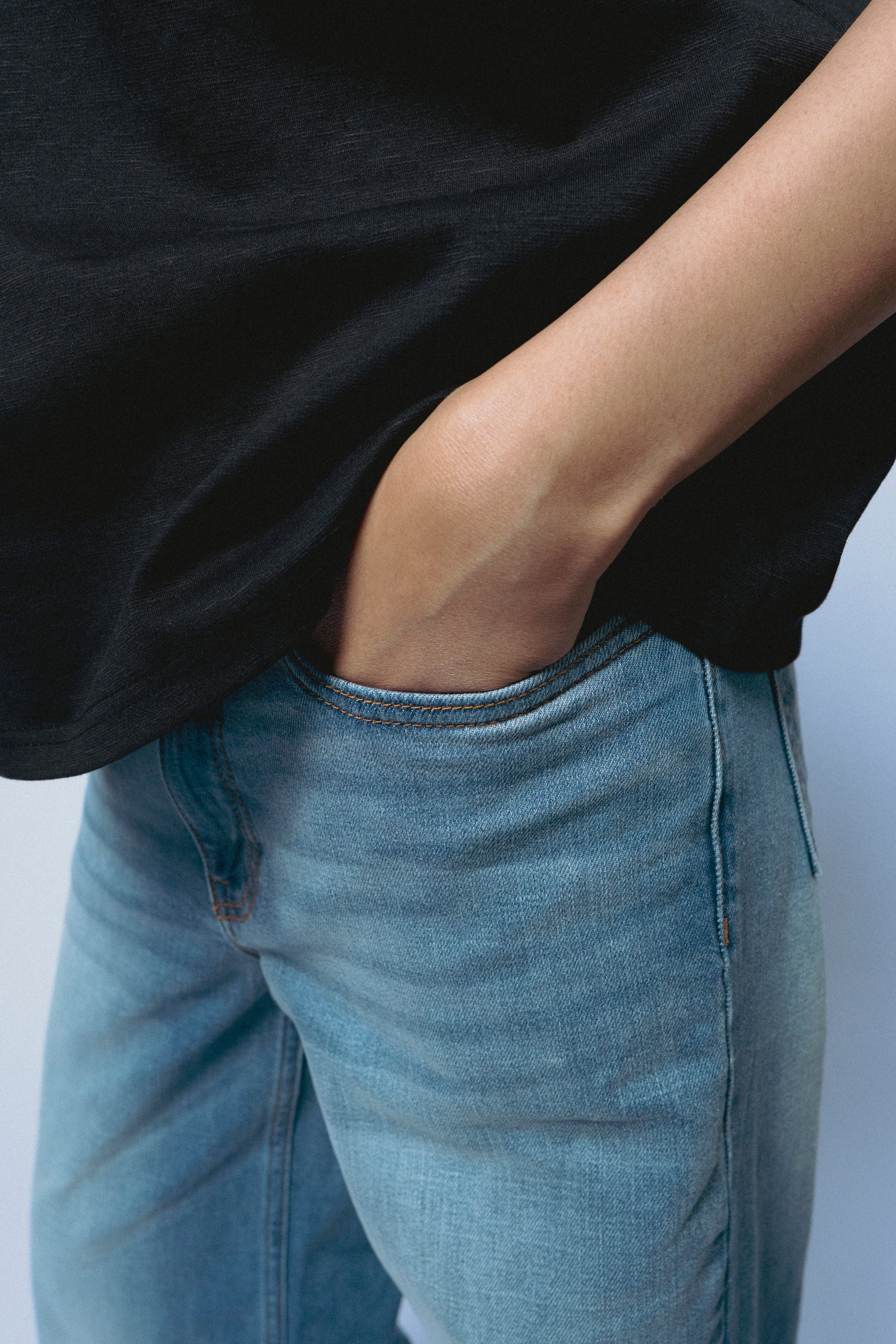 Person wearing a SOH black shirt and SOH blue jeans with hands in pockets on a light gray background