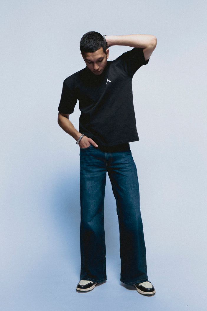 Man wearing a black SOH Mark T-shirt and blue jeans against a light blue background
