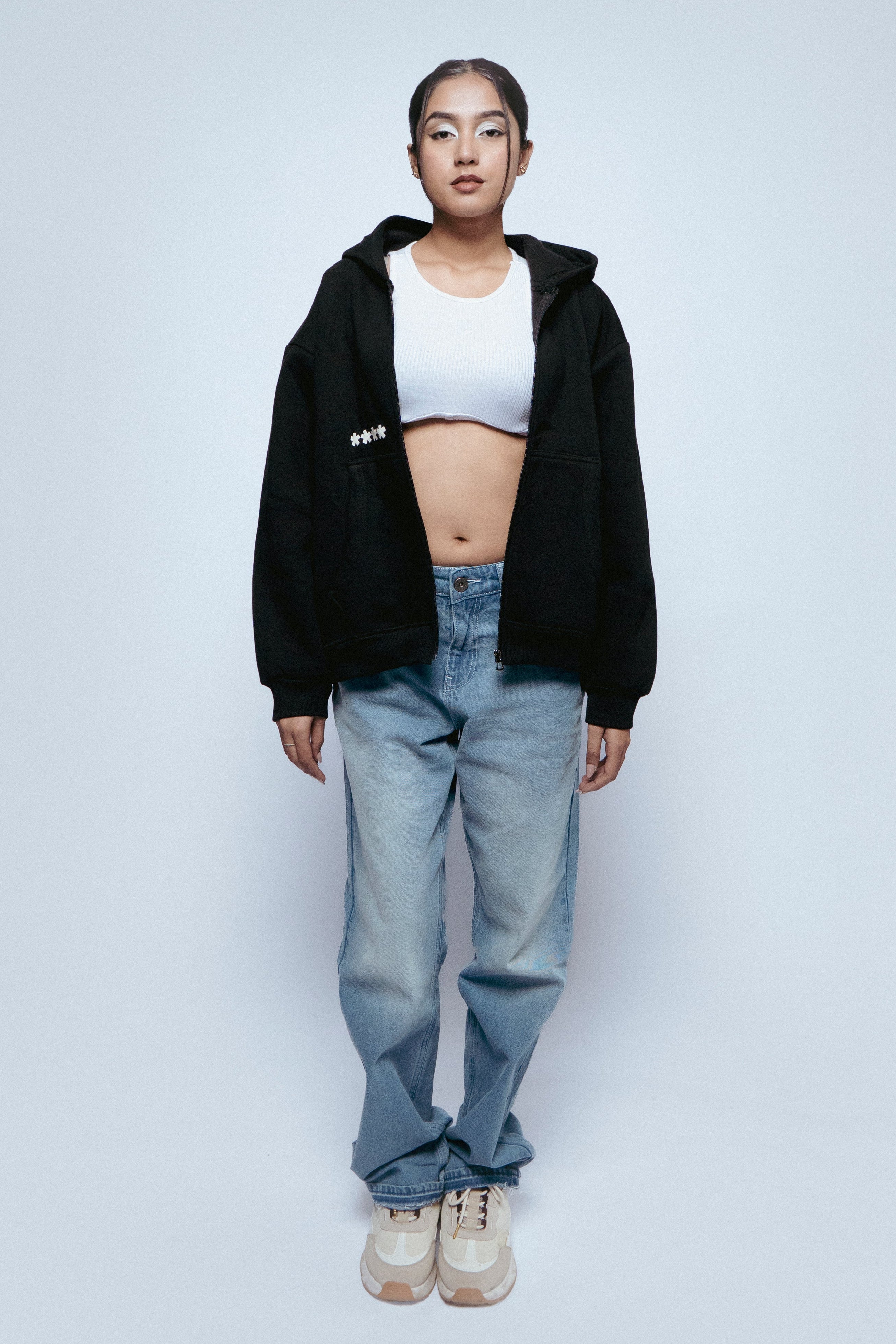 Person wearing a SOH hoodie, white crop top, SOH Noir jeans, and white sneakers on a light gray background