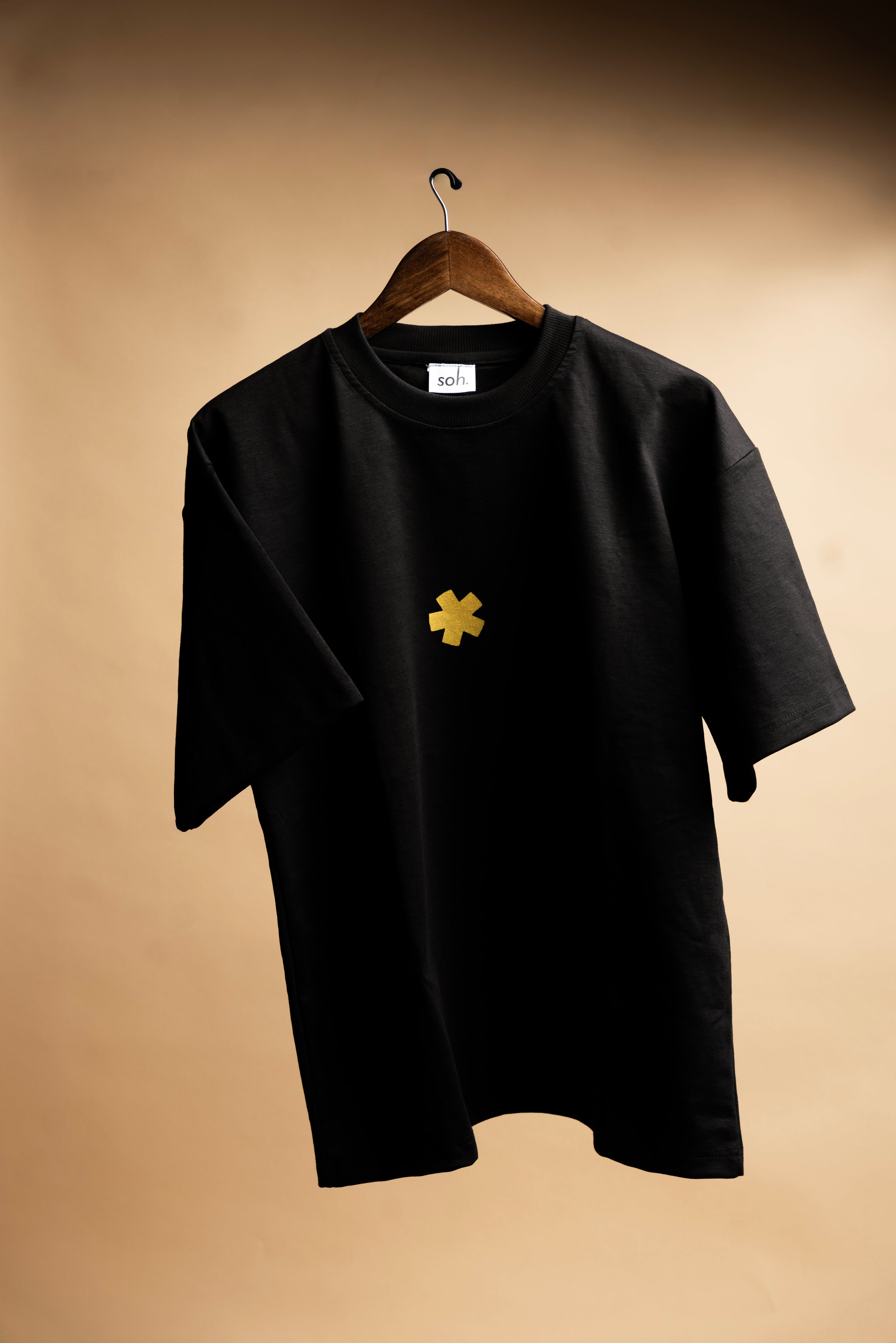 Black SOH Padel Tshirt with a yellow SOH logo design on a hanger against a beige background