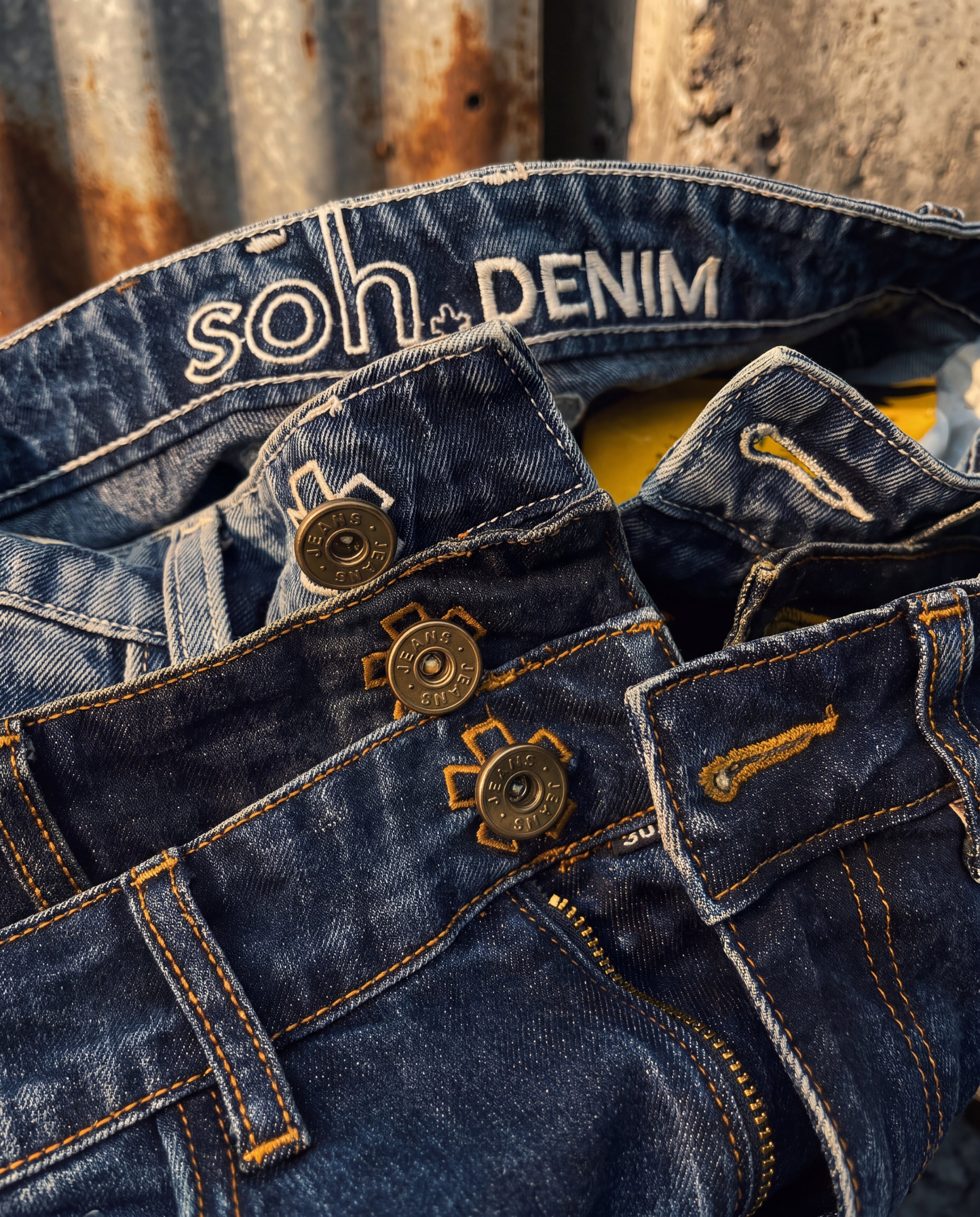 Close-up of soh dark blue denim jeans with branding on the waistband, metal cargo-style buttons, and contrast stitching, photographed against a rustic corrugated metal background.
