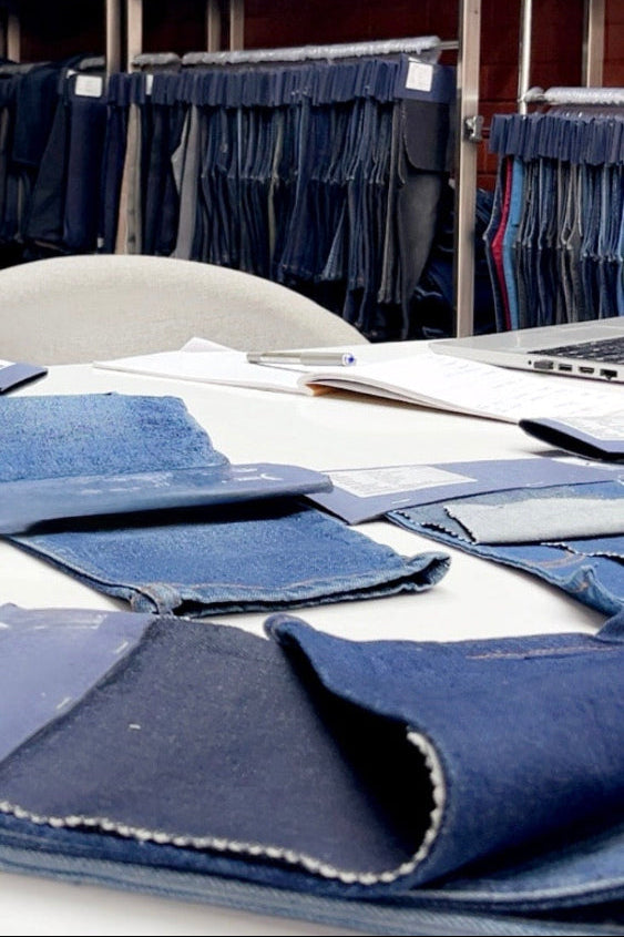 A close-up of folded and laid-out blue denim fabric swatches on a table, with racks of hanging denim garments visible in the background.