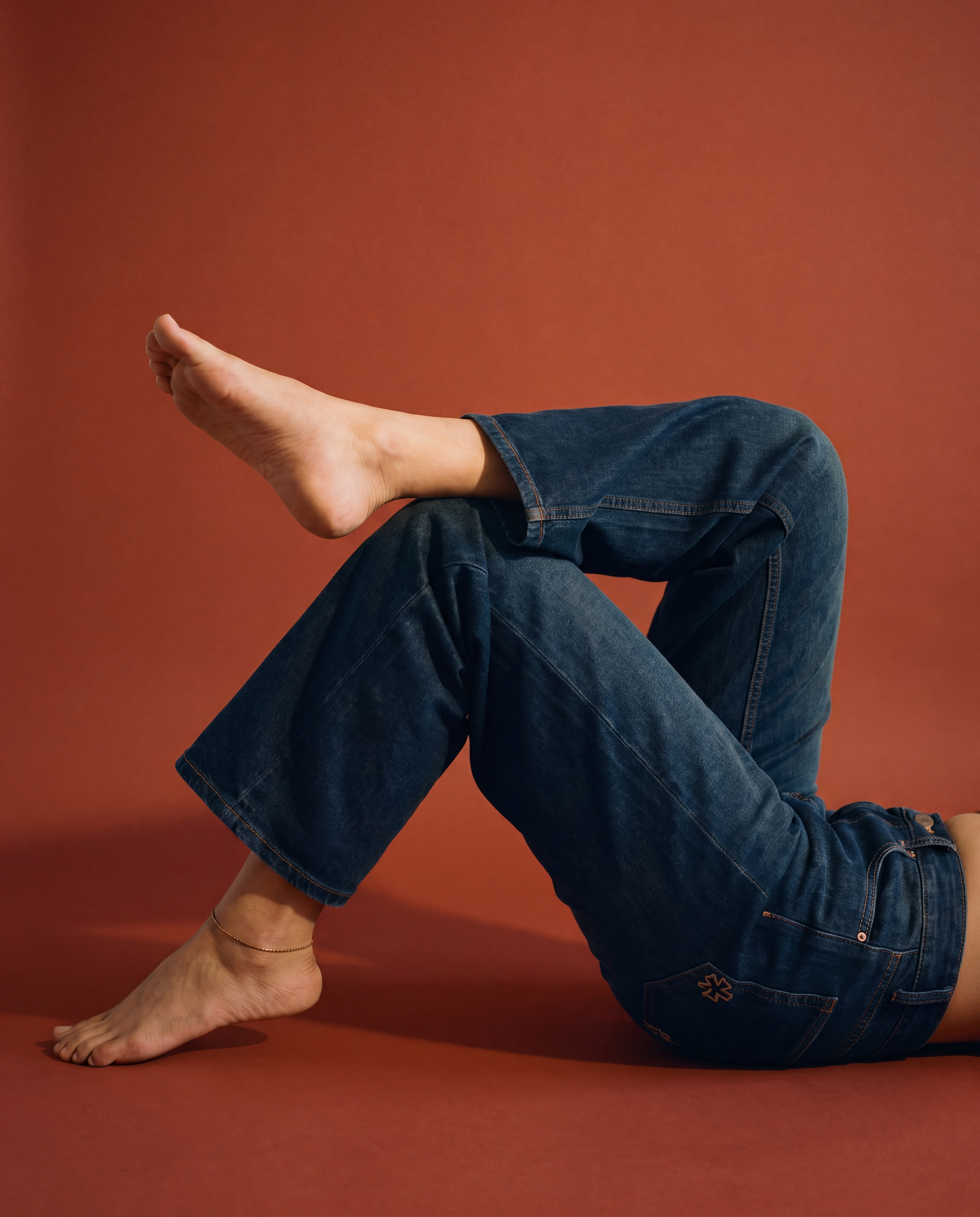 A person reclining on a warm terracotta studio floor wearing SOH's dark blue denim jeans, with one leg bent upward. 