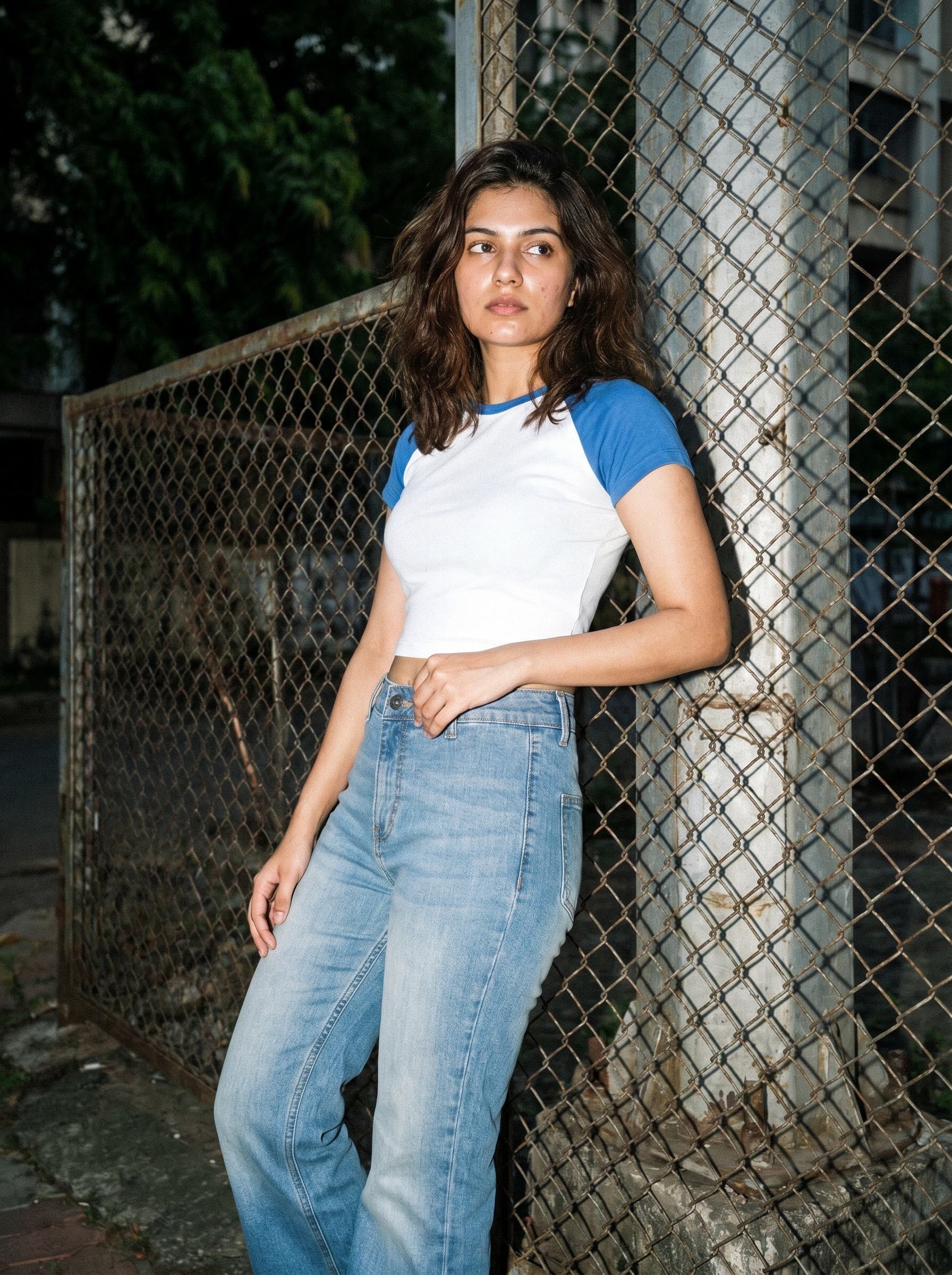 Woman wearing a white and blue shirt with jeans leaning against a chain-link fence.